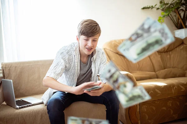 Generation z teenager wastes money by scattering it while gaming on laptop. 14-year-old boy in briquettes is drinking dollars around the apartment. The child does not know how to handle money. Financial problems in teens.