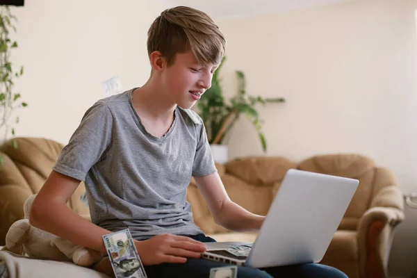 freelance teenager recounts money earned online on laptop and happy with earnings. boy studies online and earns money from comfort of home. child counting money showing it friends on conference call