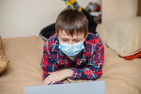 stock image teenage boy in protective medical mask coughs in fist. child remotely does lessons lying on bed near laptop. Online training during coronavirus quarantine. Home education