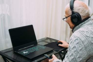 Süveter ve gözlük takan yakışıklı yaşlı adam evde çalışma masasında otururken dizüstü bilgisayar kullanıyor ve gülümsüyor. Dedem sosyal medyada torunlarıyla sohbet ediyor. Telefonu kullanan kıdemli adam.