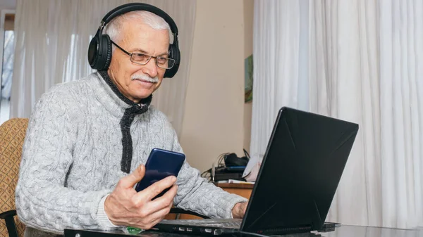 Handsome old man dressed in sweater and eyeglasses is using laptop and smiling while sitting at his work table at home. Grandpa chats in grandchildren on social media. senior man using phone.