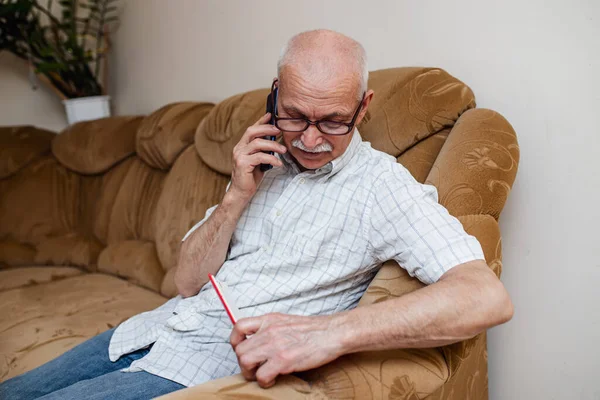 An elderly man talks on the phone and writes down the data in a diary or glider. Organization of business in retirement. A pensioner works remotely, conducts business from home.