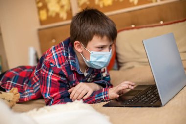 Koruyucu tıp maskeli genç bir çocuk yumruğuyla öksürüyor. Çocuk dizüstü bilgisayarın yanında yatarak uzaktan ders alıyor. Çevrimiçi alışveriş, e-öğrenme, internet konsepti üzerine uzaktan çalışma.