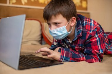 Koruyucu tıp maskeli genç bir çocuk yumruğuyla öksürüyor. Çocuk dizüstü bilgisayarın yanında yatarak uzaktan ders alıyor. Çevrimiçi alışveriş, e-öğrenme, internet konsepti üzerine uzaktan çalışma.