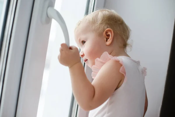 baby is trying to open the window in the apartment. No lock on the window. Dangerous home for a baby. Fall safety for children. The child stands on the windowsill of a closed window and tries to open the window sash without a lock