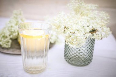 Yazlık terasta şeffaf bir bardak, bir dilim limon ve bir buket mürver. Ambucus şurubuyla sulu içecek. Çiçek açan bir ağaçtan tıbbi bir ürün.