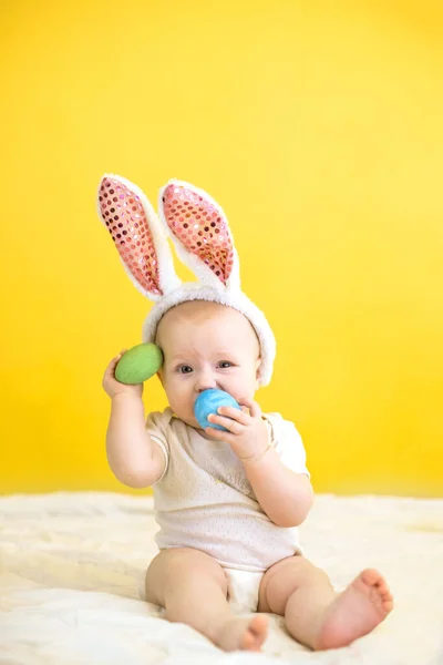 funny child gnaws toy Easter egg on yellow background. Toddler, wearing rabbit hat, curled up funny face. Easter concept.