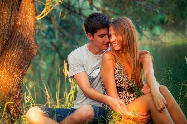 一对男女青少年牵着手 男人爱那个女孩 年轻的恋人坐在湖边靠近树的地方 — 图库照片