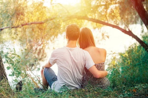 カップルの男の子と女の子のティーンエイジャーの手を握って 男はその少女を愛している 若い恋人たちは木の近くの湖の岸に座っている — ストック写真