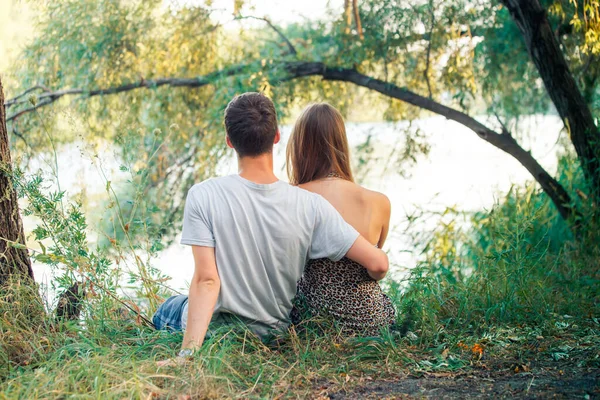 カップルの男の子と女の子のティーンエイジャーの手を握って 男はその少女を愛している 若い恋人たちは木の近くの湖の岸に座っている — ストック写真