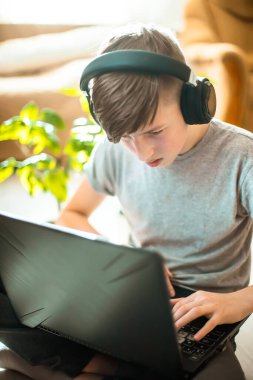 Diş teli takan bir genç dizüstü bilgisayarda yerde oturuyor. Kış bahçesinden gelen yeşil bitkilerin yanında şık iç mekanlarda çalışan çocuk evde online ders veriyor.