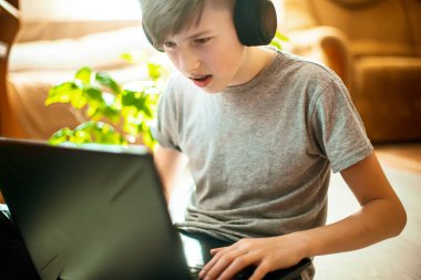 Diş teli takan bir genç dizüstü bilgisayarda yerde oturuyor. Kış bahçesinden gelen yeşil bitkilerin yanında şık iç mekanlarda çalışan çocuk evde online ders veriyor.