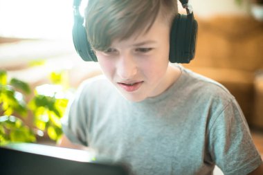 Diş teli takan bir genç dizüstü bilgisayarda yerde oturuyor. Kış bahçesinden gelen yeşil bitkilerin yanında şık iç mekanlarda çalışan çocuk evde online ders veriyor.