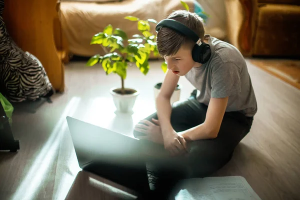 stock image teenager in braces on teeth programs sitting at laptop on floor. boy in fashionable interior near trendy green plants from winter garden teaches lessons online at home