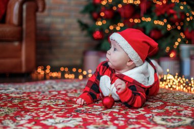 Noel Baba gibi giyinmiş altı aylık bir bebek. Yılbaşı kartı için arka plan. Çocuk aydınlık çelenklerin ve bir Noel ağacının arkaplanındaki yazıtlara bakıyor. Boşluğu kopyala