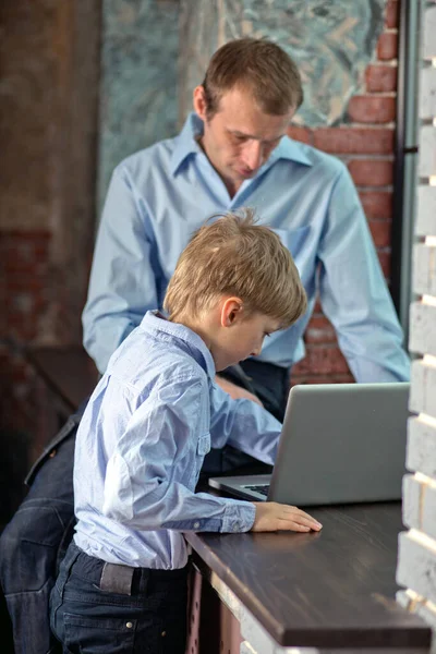 Dad sits on windowsill teaches son to work on computer. Father and son playing on laptop. Father is looking for gift to son in online store in modern loft. Vertical, blurred background.