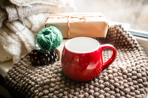 An empty red Christmas cup on a cozy blanket on the windowsill near the window. Gift wrapping with natural string and recycled paper.