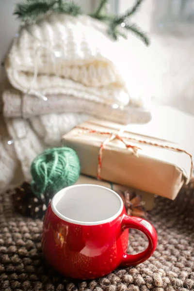 A cozy evening at home for wrapping gifts for Christmas. stack of cozy white sweaters on the windowsill by the window. Preparation for tea drinking. A red empty cup on a knitted napkin