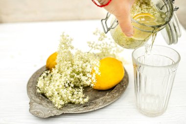 Yazlık terasta şeffaf bir bardak, bir dilim limon ve bir buket mürver. Ambucus şurubuyla sulu içecek. Çiçek açan bir ağaçtan tıbbi bir ürün.