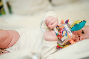 Yeni doğmuş bir bebek yan yatar ve eğitici bir oyuncağı elinde tutar. Dokunsal duyular için oyuncaklar ve bebeklerin şekilleri ve renkleri üzerine çalışma. Odaklanmamış