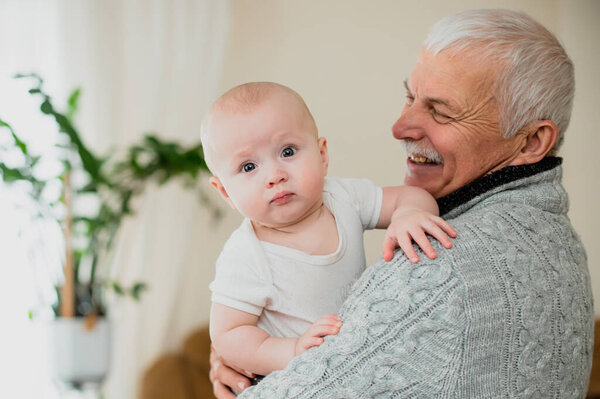 A little girl peeks out from over her grandfather's shoulder. An old man plays at home with grandson. A gray-haired elderly man with a mustache holds a girl of 6 months in arms.