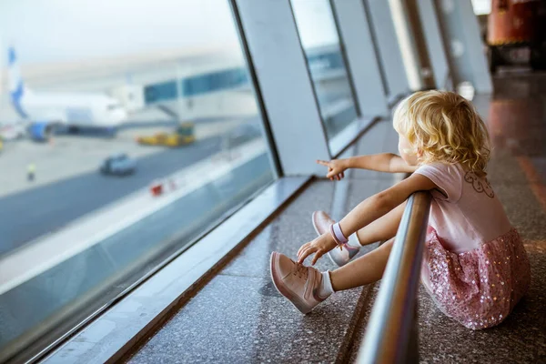 little girl sits by window at airport and points finger at plane. child in airport departure lounge. Girl sitting on floor waiting to board plane.