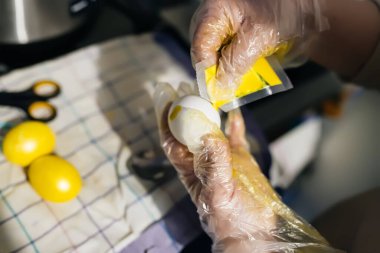 Painting eggs for Easter with artificial dyes in yellow. Hands in polyethylene gloves hold a chicken egg covering with paint from sachet. Part of the series Easter preparation at home