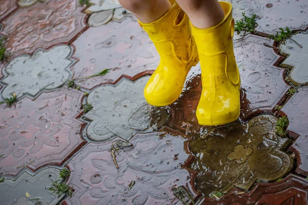 A child in yellow rubber boots during the rain gallops through puddles on concrete tiles near the house