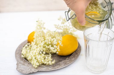 Hand pouring a refreshing summer drink of elderberry and lemon into a faceted glass on a white table.