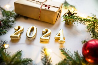 Beyaz zemin üzerinde altın rakamlar 2024, el işi hediyesi, Noel ağacı dalları ve kırmızı süslemeler ve yılbaşı ışıkları.