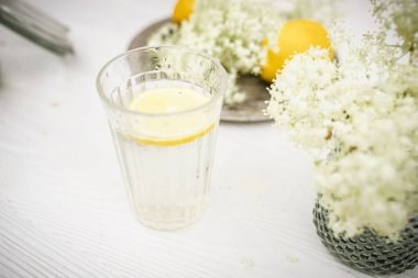 Beyaz arka planda taze mürver çiçekleri olan vazonun yanında bir dilim limon bulunan bir cam.