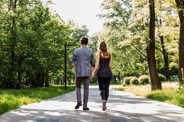 Güneşli bir günde şehir parkında yürüyen genç bir çiftin arka görüntüsü.