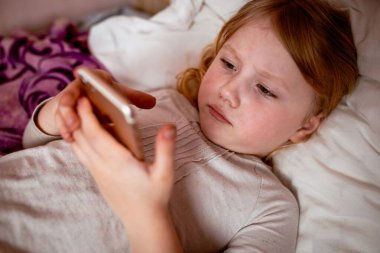 Küçük kız yatakta uzanıp cep telefonuna bakıyor.