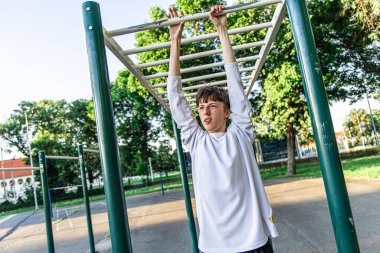 Genç bir adam açık hava spor salonunda maymun parmaklıklarından sarkarken görülüyor. O, uzun kollu beyaz bir gömlek giyiyor. Parmaklıklar metalden yapılmış ve yeşile boyanmış. Parmaklıkların arkasında yeşil çimenlik bir alan var. Parlak güneşli havadaki antrenmanına odaklanmış.