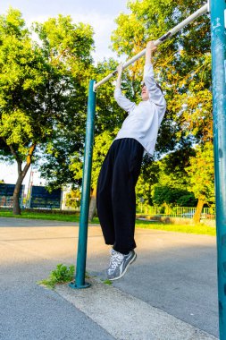 Beyaz gömlekli ve siyah pantolonlu genç bir adam parktaki bir barfiks barında asılı duruyor. O, kararlı bir ifadeyle gökyüzüne bakıyor. Güneş parlıyor ve arkadaki ağaçlar yeşil ve yemyeşil..