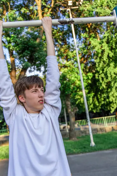 Uzun kollu beyaz gömlekli genç bir çocuk parktaki bir barfiks barında sallanıyor. O, yüzünde odaklanmış bir ifadeyle bara bakıyor. Arka plan yemyeşil ağaçlar ve parlak mavi gökyüzü ile dolu..