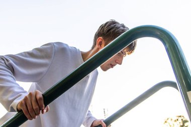 Uzun kollu beyaz gömlek giyen kısa kahverengi saçlı genç bir adam açık havada metal merdivenlerden yukarı çıkıyor. O tırabzanı kavrayıp yukarı doğru giderken, güneş arka planda parlıyor..