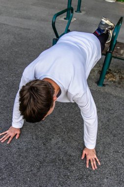 Beyaz gömlekli genç bir adam yeşil bir açık hava egzersiz bankında şınav çekiyor. Spor ayakkabıları giyiyor ve antrenmanına odaklanmış durumda. Toprak gri asfalt..