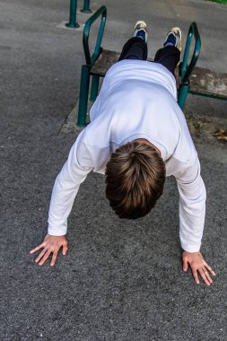 Bir insan dışarıda metal bir spor aletinde şınav çekiyor. Beyaz gömlek ve siyah pantolon giyiyorlar. Zemin asfalt..
