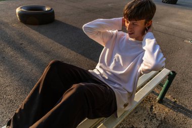 Genç bir adam antrenman sırasında dışarıda bir bankta mekik çekiyor. Uzun kollu beyaz bir gömlek ve koyu renk pantolon giyiyor. Arka planda bir lastik var. Güneş arka planda batıyor..