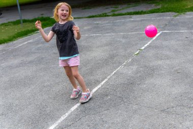 Pembe voleybolu ileri geri fırlatırken küçük kız kahkahaları havayı doldurdu. Sarı saçlı genç bir kız kaldırımlı oyun sahasında pembe bir topla oynuyor..