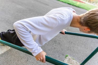 Uzun kollu beyaz gömlek ve siyah pantolon giyen genç bir adam açık hava spor salonunda paralel barlara daldırıyor. Vücudu dirseklerinden bükülmüş ve yere doğru eğiliyor. Gücünü ve dikkatini gösteriyor..