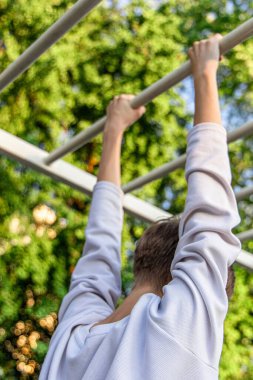 Bir kişi aşağıdan izlenen oyun parkındaki bir dizi yatay çubuğa asılır. Beyaz bir sweatshirt giyiyorlar ve sırtları kameraya dönük. Arka planda büyük yeşil bir ağaç var..