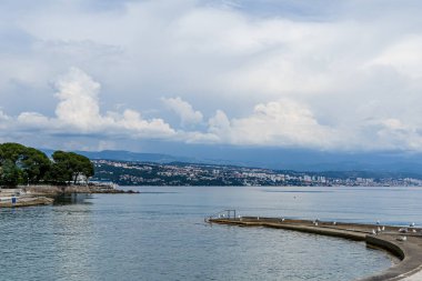 Hırvatistan 'ın Opatija kentindeki Adriyatik Denizi manzarası, suya uzanan küçük kayalık bir kıyı şeridi. Beyaz kabarık bulutlar mavi bir gökyüzünde sürükleniyor ve şehir kıyı şeridi boyunca görülebiliyor. Martılar kayalık sahilde dinleniyor..