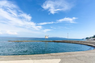 Hırvatistan 'ın Opatija kıyılarından güzel bir manzara, tüylü beyaz bulutlarla kaplı açık mavi bir gökyüzünü gösteriyor. Sakin turkuaz deniz ufka doğru uzanır ve arka planda uzak bir dağ sırası görülebilir..