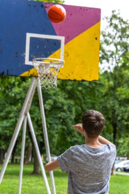 Genç bir adam dışarıda basketbol sahasında basket oynuyor. O gri bir tişört giyiyor ve topa doğru kavis çizerken bakıyor. Basketbol potası mavi, sarı ve pembe renklerle boyanmıştır..