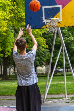 Genç bir adam bir basketbol topunu açık hava sahasında potaya doğru fırlatırken görülüyor. Basketbol topu havada ve oyuncu sağ eliyle elini uzatıyor. Saray yeşil ağaçlar ve çimlerle çevrili..