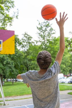 Bir genç gün boyunca açık hava sahasında basket potasına doğru basket atıyor. Basketbol topu havada ve çocuk da atış hareketinin tam ortasında. Saray yemyeşil ağaçlar ve çimlerle çevrili..