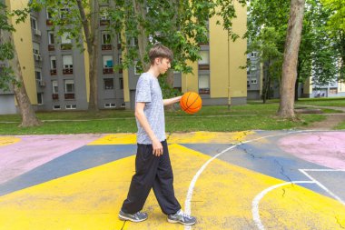 Gri tişörtlü ve siyah pantolonlu genç bir adam dışarıda basket topu sürüyor. Mahkeme sarı, mavi ve pembe çizgilerle boyanmıştır. Adam görüntünün sağ tarafına doğru yürüyor. Arka planda bir bina kompleksi var..