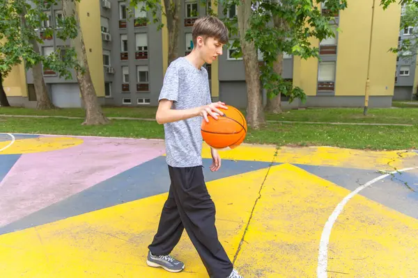 stock image A young man wearing a gray t-shirt and black pants is dribbling a basketball on an outdoor basketball court. The court is painted with yellow, pink, and blue lines. The man is walking toward the right side of the image, looking down at the ball.